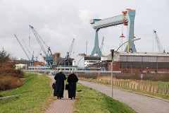 Passanten auf dem Deich in Hamburg Neuenfelde - Portalkran auf dem Werftgelände der Sietas Werft.