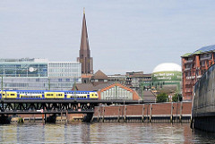 Ein Zug fährt über die Oberhafenbrücke - daneben das ehem. Marktgebäude in dem sich jetzt das Haus der Fotografie befindet. Im Hintergrund der Turm der Hamburger St. Jacobikirche.