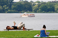 Entspannung am Ufer der Aussenalster - Ruhe vom Grossstadttrubel. Eine Frau liegt auf der Bank und liest ein Buch, eine andere sitzt auf der Wiese mit ihrem Labtop - im Hintergrund das Alstercabrio .