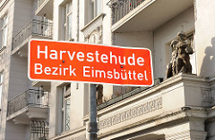 Stadtteilschild Harvestehude; rotes Schild, weisse Schrift - Putten auf Balkonsims.