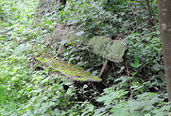 Ruhebank aus Holz - vermoderte Sitzgelegenheit am Rande des Wanderwegs durch das Hamburger Naturschutzgebiet Raakmoor - Grünpflanzen überwuchern die Holzbank, die mit Moos bedeckt ist.