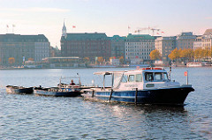 Motorboot mit Kähnen im Schlepp auf der Hamburger Binnenalster - im Hintergrund der Hamburger Jungfernstieg und das Alsterpavillon.