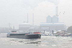 Das Binnenschiff NIEDERSACHSEN fährt durch das eisbedeckte Wasser der Billwerder Bucht - das Schiff hat Kohle für das Kraftwerk Tiefstack gebracht, das im Hintergrund im Dunst zu erkennen ist.