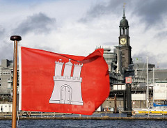 Die Flagge der Hansestadt Hamburg weht im  Wind - im Hintergrund der Turm des Hamburger Wahrzeichens, die St. Michaeliskirche / Michel.