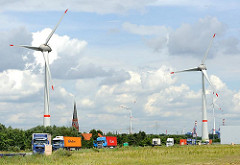 Gewerbegebiet Hamburg Altenwerder, geparkte Lastkraftwagen mit Containern beladen - Windkraftanlagen mit riesigen Rotorblättern - dazwischen der Kirchturm der St. Gertrudkirche.