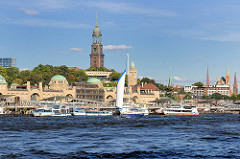 Blick über das Wasser der Elbe zu den St. Pauli Landungsbrücken - Fahrgastschiffe / Hafenrundfahrt liegen am Ponton, ein Segelschiff kreuzt auf der Elbe; im Hintergrund Kirchtürme der Hansestadt - St. Michaeliskirche; Michel in der Bildmitte.