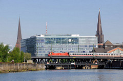 Blick vom Oberhafenkanal zur Oberhafenbrücke - ein Zug mit roter Lokomotive überquert die Brücke über den Kanal - im Hintergrund das Deichtorcenter und die Kirchtürme der St. Petri und St. Jacobikirche.
