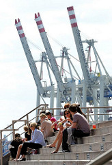 Mittagspause am Hafen - Angestellte der umliegenden Bürohäuser geniessen die Sonne an der Grossen Elbstrasse in Hamburg Altona; im Hintergrund hochgefahrene Containerkräne des HHLA Terminals Tollerort.