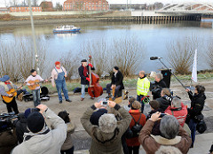 Musiker am Ufer des Spreehafen spielen auf der Veranstaltung zum Abriss des Zollzauns. Zuschauer fotografieren die Veranstaltung vom Deich - im Hintergrund ein Polizeiboot der Hamburger Wasserschutzpolizei im Spreehafen.