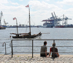 Blick von der Grossen Elbstrasse auf den Hamburger Hafen - ein Pärchen sitze auf der Kaimauer am Wasser des Hamburger Flusses, ein historisches Segelschiff fährt stromabwärts. Im Hintergrund die Terminalanlagen von Tollerort.