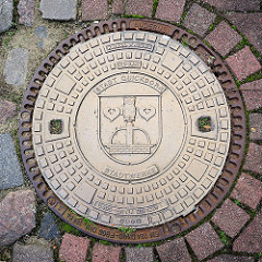 Gusseiserner Gullideckel der Stadt Quickborn / Stadtwerke; Wappen der Stadt, Brunnen mit Eule, zwei Seeblätter.