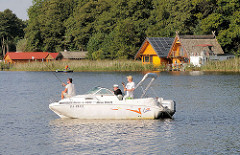 Angelboot und Bootshäuser am Mirower See - Hausdächer hinter Schilf am Seeufer.