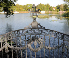 Gusseisernes Dekor, Zierelement an der Brücke der Mirower Schlossinsel - im Hintergrund Bootshäuser am Ufer des Mirower Sees.