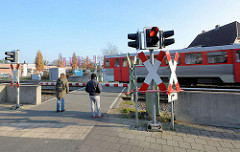 Fussgängerüberweg mit geschlossener Schranke und rotem Stopplicht am Bahnhof Quickborn - ein Zug der AKN fährt vorbei.
