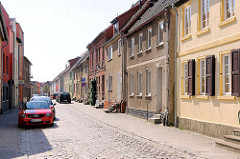 Einstöckige Wohnhäuse mit Rauhputz oder farbiger Fassade, parkende KFZ / Kopfsteinpflaster.