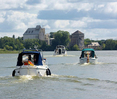Motorboote überqueren den Röblinsee und folgen dem Lauf der Havel in Fürstenberg; im Hintergrund Industriegebäude einer verlassenen Fabrik am Seeufer.