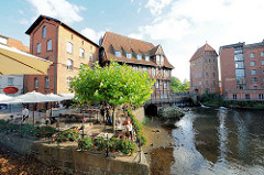 Restaurant / Café im Freien am Fischmarkt von Lüneburg - Gäste sitzen unter Sonnenschirmen an der Ilmenau. Im Hintergrund das Gebäude der Lüner Mühle / Alte Ratsmühle aus dem Jahr 1597; Ilmenau Hafen.