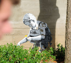 Graffiti an einer Hauswand in Lüneburg - ein Kind / kleines Mädchen sitzt im Gras + pflückt Blumen - es wird von einem Mann beobachtet.