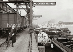 Schuten liegen am Afrikakai des Hamburger Südwesthafens - einige der Lastkähne sind leer, während ein anderer mit Holzkisten hoch beladen ist. Unter den Halbportalkranen steht ein Güterzug; im Hintergrund dampft ein Schlepper in dem Hafenbecken.