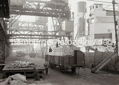 Mit Holzbohlen beladene Güterwaggons sind am Kai auf den Gleisen vor den Frachter geschoben worden - mit dem Kran werden die Bündel Holzbretter von den offenen Waggons gehoben und auf das Schiff transportiert.