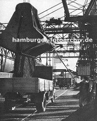 Kohlekai am Altonaer Hafen; Kräne schütten die Kohle in die bereitstehenden Lastwagen -  Hafenarbeiter dirigieren das Schüttgut mit einer langen Stange - im Hintergrund ein Güterwaggon auf den Schienen der Hamburger Hafenbahn; darüber die Förderan