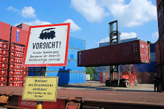 Ein Schwerlaststabler / Gabelstabler transportiert einen Container vom Containerlager zum Güterzug. Schild Vorsicht privater Bahnübergang.