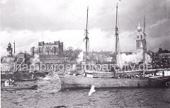Ein Dreimaster wird von einem Schlepper aus dem Hamburger Hafen geschleppt. Am Elbufer links der Uhrturm von den St. Pauli Landungsbrücken, daneben der Eingang von der Hochbahnstation Landungsbrücken sowie das Gebäude der Hamburger Seewarte.