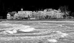 Wellen und Gischt - windiger / stürmischer Tag an der Ostsee - im Hintergrund die Strandpromenade mit Gebäuden.