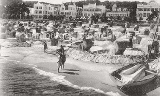 Ostseestrand in Binz auf Rügen - historisches Foto um 1900; Holzboote / Ruderboote im Verleih - Damen in Kleidern mit Hüten sitzen in Strandkörben; Villen in Bäderarchitektur / Bäderstil an der Strandpromenade.