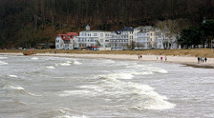 Wellen und Gischt - windiger / stürmischer Tag an der Ostsee - im Hintergrund die Strandpromenade mit Gebäuden.