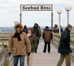 Seebrücke - Schild Seebad Binz, Urlauber mit warmen Jacken und Mützen - windiger Frühlingstag.