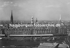 Blick über den Sandtorhafen und dem Sandtorkai zur Hamburger Speicherstadt mit ihren Giebeln und Türmen der Backsteinarchitektur. Schiffe liegen am Kai und werden entladen und die Ladung in den Lagerschuppen am Kaispeicher verstaut.