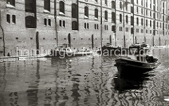 Barkassen liegen an der Mauer eines Backstein-Speichers in der Speicherstadt; die Mauern der  Speicherge- bäude sind mit Holzstämmen gegen Beschädigungen durch Schiffe geschützt. Aus einigen Luken hängen Strickleitern.