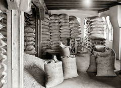 Der Kaffee aus Kolumbien ist in Jutesäcke vernäht und bis an die Decke des Speicherbodens gestapelt. Die Säcke tragen die Aufschrift Produkt of Colombia - 70 Kilos. Ein Haufen Kaffee ist auf dem Lagerboden ausgeschüttet.