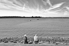 Elbufer der Unterelbe bei Wedel - ein älteres Paar sitzt auf der Uferbefestigung und blickt auf die Elbe - Schwarz Weiss Aufnahme.