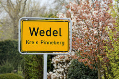 Ortsschild / Stadtschild Wedel, Kreis Pinneberg - im Hintergrund blühende Sträucher.