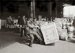 Drei Lagerarbeiter stemmen eine schwere Holzkiste auf eine Sackkarre. Im Hintergrund stehen weitere Kisten und Holzfässer im Kaischuppen und auf der Laderampe. Arbeiter arbeiten mit Handkarren im Lagerschuppen; ca. 1934.