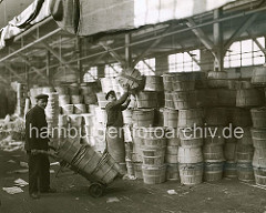 Die Arbeiter im Fruchtschuppen A am Baakenhafen, eines der vielen Hafenbecken und Kaianlagen des Hamburger Hafens, transportieren die Fässer mit Sackkarren vom Hafenkai zum Stauraum. Dort wird die empfindlichen Fracht von Hand gestapelt; ca. 1930