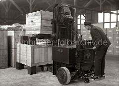 Vorsichtig stapelt ein Arbeiter mit dem Elektrohubwagen zwei Holzkisten übereinander. Die Kisten stehen auf Blöcken als Abstandshalter, damit die Gabel des AEG Elektrowagens dazwischen gefahren werden kann; ca. 1934.