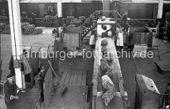 Die Bananenstauden werden von ihrem Lagerplatz im Kaischuppen auf einem Förderband zur Laderampe transportiert, wo die Südfrüchte in einen bereit stehenden Güterzug verladen werden. Vor der Verladung wird die Qualität der Ware geprüft.