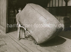 Ein großer Ballen wird von einem Hafenarbeiter von Hand mit Hilfe einer Sackkarre in den Kaischuppen transportiert; ca. 1934.