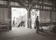 Hafenarbeiter transportieren Kisten mit der Sackkarre auf der Laderampe und im Lagerschuppen. Im Hintergrund liegt ein Frachtschiff am Kai; ca. 1932.