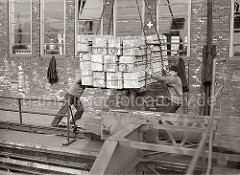 Ein Kranführer setzt die Hieve Apfelsinenkisten auf der Galerie des Fruchtschuppens ab - Lagerarbeiter dirigieren die Ladung an ihren Platz; im Vordergrund das Schienensystem mit Führung des Halbportalkrans; ca. 1930.