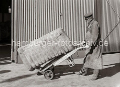 Der Kaiarbeiter transportiert einen Ballen, der mit Bandeisen zusammengehalten wird auf seiner Sackkarre. Der Hafenarbeiter trägt seine Dienstmütze und eine Schürze zum Schutz seiner Kleidung; ca. 1934.