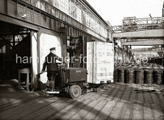 Ein Lagerarbeiter hat eine große Holzkiste auf die Gabel des Elektrohubwagen geladen und fährt sein Transportgut auf die Lagerrampe des Schuppens 83 am Chilekai des Oderhafens. Auf der Rampe stehen Metallfässer.