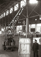 Eine große Holzkiste wird in einer der vielen Lagerhallen des Hamburger Hafens mit einem Laufkran transportiert. Vier Metallkrallen, die sich an der Eisenkette des Kranhakens befinden, tragen die Kiste.