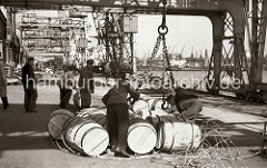 Hafenarbeiter schliessen das Transportnetz um eine Hieve Fässer, dann haken sie das Netz in die Kranhaken ein und die Ladung wird an Bord des Frachtschiffs gebracht. Im Hintergrund rollen Arbeiter die Holzfässer von Hand auf die Rampe.