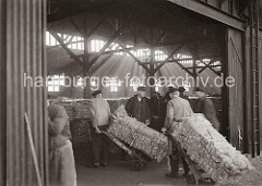 Kaiarbeiter transportieren dei Wollballen mit Sackkarren von der Laderampe in den Kaischuppen. Durch das Oberlicht des Lagerhauses fällt das Licht auf den mit Ballen gefüllten grossen Raum; ca. 1934.