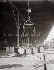 Vier Fässer hängen im Ladegeschirr des Hafenkrans über der Laderampe des Lagerschuppens. Im Hintergrund steht ein Lastwagen an der Rampe; ca. 1930.
