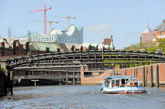 Eine Barkasse der Hamburger Hafenrundfahrt / Speicherstadtrundfahrt fährt auf dem Zollkanal unter der Jungfernbrücke - im Hintergrund die Kibbelstegbrücke und Kupferdächer der Speicherstadt sowie die Baustelle der Elbphilharmonie.